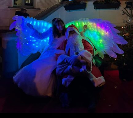 Weihnachtsengel in Rackwitz Eine Person in einem roten Kostüm wie Santa Claus, umgeben von leuchtenden Flügeln und einer Frau.